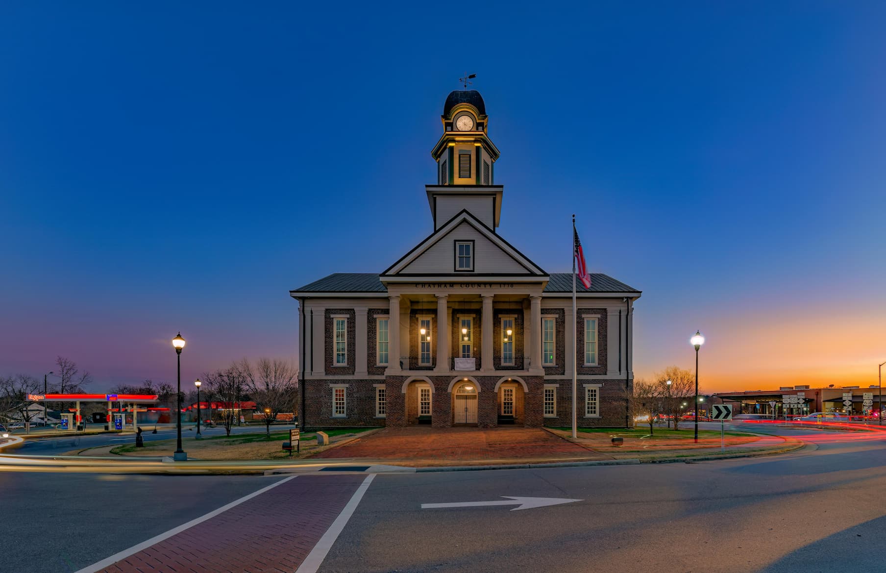The historic Chatham County Courthouse illuminated at twilight, featuring a grand brick facade with white columns, a distinctive clock tower with golden accents, and an American flag. The building is captured against a deep blue evening sky with orange sunset hues along the horizon, while streetlights and traffic create light trails around the circular court square.
