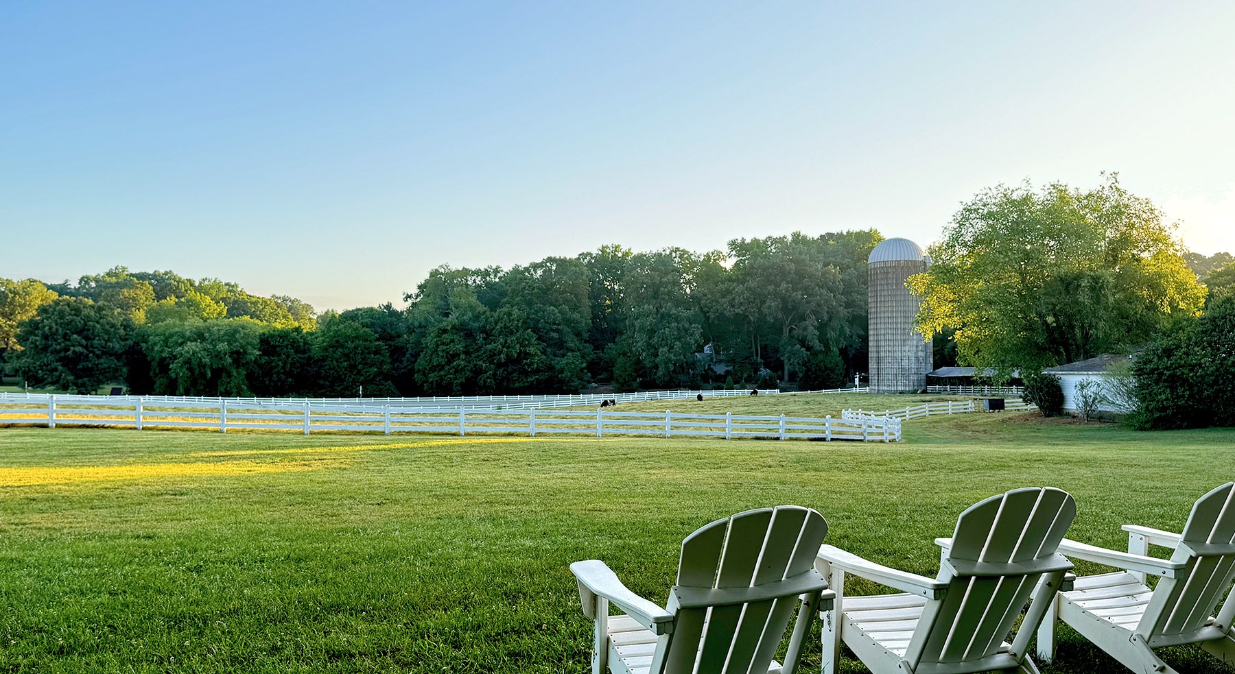 A serene rural landscape at Fearrington Village featuring a well-maintained pasture enclosed by white fencing. In the foreground are several white Adirondack chairs on a lush green lawn. Beyond the fence is a pastoral scene with a tall cylindrical silo visible on the right side, surrounded by mature trees. The scene is bathed in golden evening light under a clear blue sky, creating a peaceful countryside atmosphere.
