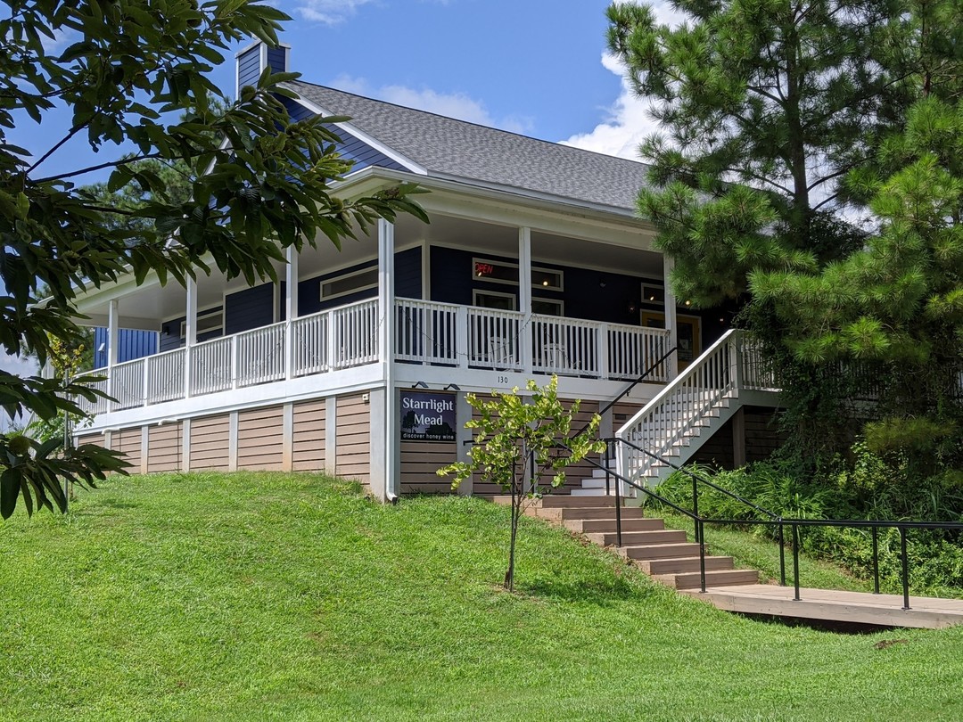 The Starrlight Mead tasting room, a charming elevated building with navy blue and tan exterior, featuring a wide covered porch with white railings and exterior staircase. The facility is nestled among lush greenery with pine trees to the right, set on a well-maintained grassy slope under a bright blue sky.