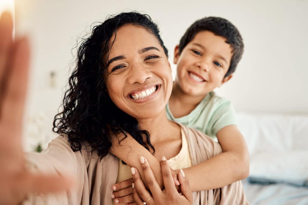 A woman smiles while taking a selfie, with a young boy hugging her from behind. Both are looking at the camera and appear cheerful in a bright indoor setting.
