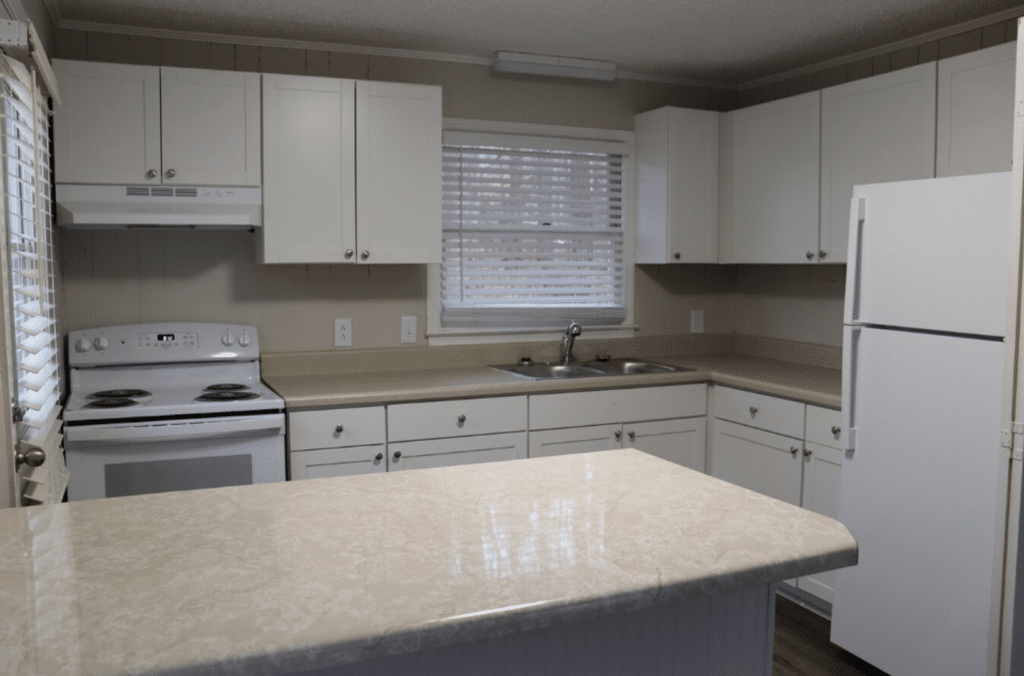 A kitchen with white cabinets, beige countertops, an electric stove, double sink, refrigerator, and window with blinds above the sink.