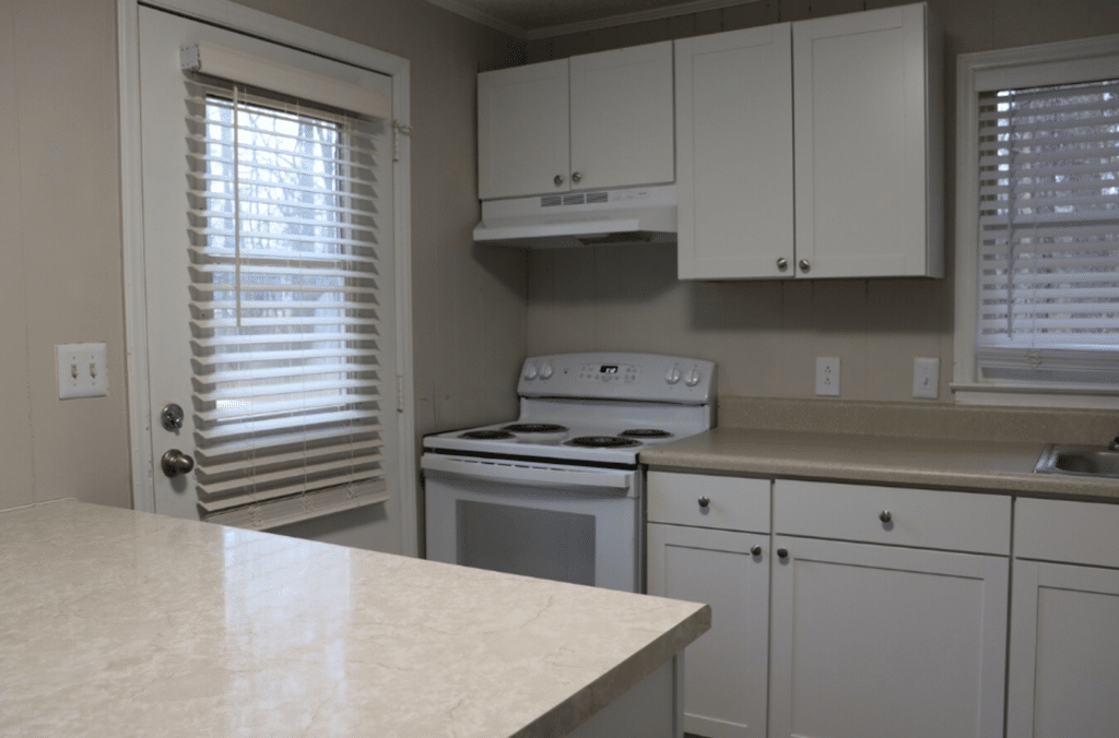 A small kitchen with white cabinets, an electric stove, a range hood, a marble countertop, and two windows with blinds.
