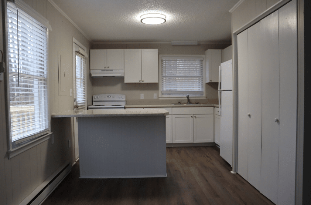 Small kitchen with white cabinets and appliances, an island counter, wood-style flooring, and windows with blinds letting in natural light.