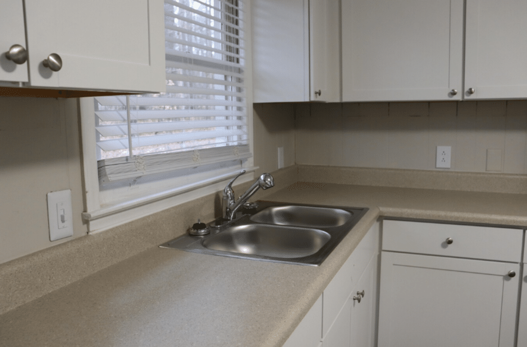 A clean kitchen countertop with a double stainless steel sink, white cabinets, and a window with blinds partially open.