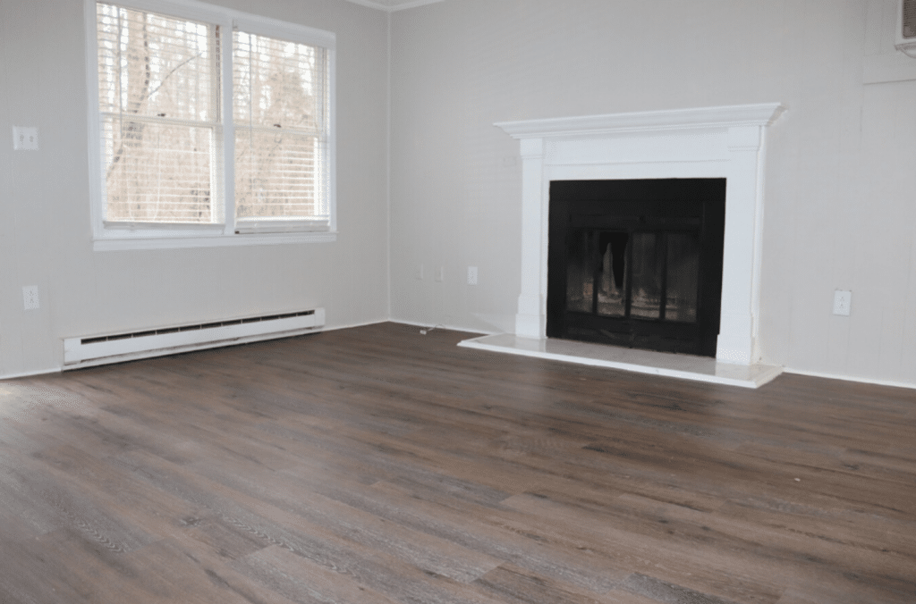 A room with dark wood flooring, a white fireplace, light-colored walls, and a window with closed blinds letting in daylight.