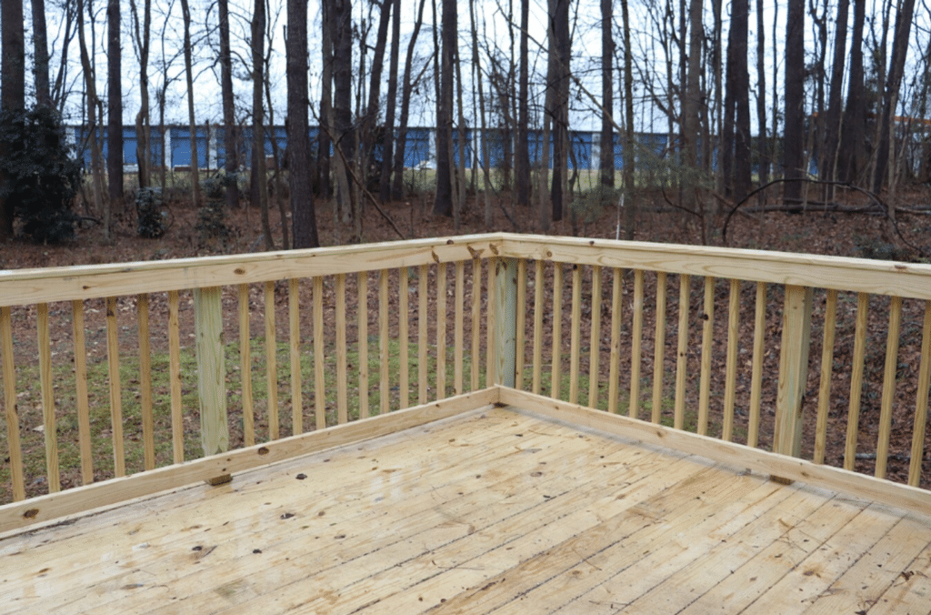A wooden deck with a railing is shown in the foreground, overlooking a yard with bare trees and a blue fence or barrier in the background.